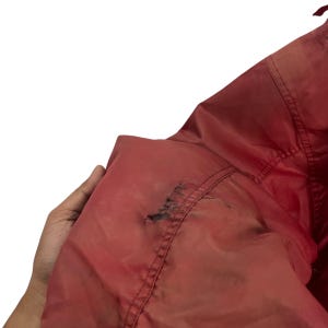 K&ouml;nnte beinhalten: Nahaufnahme einer roten Jacke mit sichtbaren Gebrauchsspuren. Der Stoff der Jacke weist Besch&auml;digungen auf, darunter ein Riss und Verf&auml;rbungen. Die Jacke wird von einer Hand gehalten, wodurch die Textur und der Zustand des Kleidungsst&uuml;cks hervorgehoben werden.