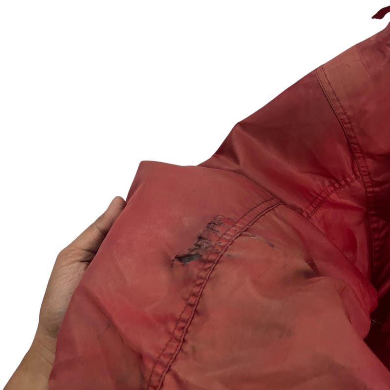 K&ouml;nnte beinhalten: Nahaufnahme einer roten Jacke mit sichtbaren Gebrauchsspuren. Der Stoff der Jacke weist Besch&auml;digungen auf, darunter ein Riss und Verf&auml;rbungen. Die Jacke wird von einer Hand gehalten, wodurch die Textur und der Zustand des Kleidungsst&uuml;cks hervorgehoben werden.
