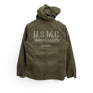 May include: Olive green military-style jacket with a hood. The back of the jacket features the text "U.S.M.C. MARINE CORPS" in a stencil font. The jacket is from Avirex Ltd.