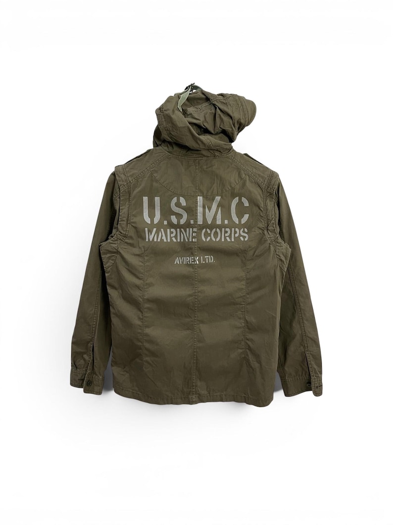 May include: Olive green military-style jacket with a hood. The back of the jacket features the text "U.S.M.C. MARINE CORPS" in a stencil font. The jacket is from Avirex Ltd.