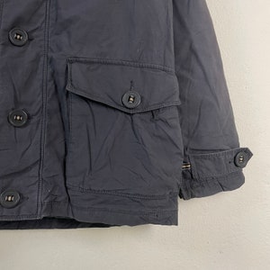 Pode incluir: Close-up de um casaco azul marinho com bot&otilde;es grandes e redondos e dois bolsos frontais. O casaco tem um punho abotoado e um design cl&aacute;ssico e utilit&aacute;rio. O tecido parece ser um material dur&aacute;vel e ligeiramente texturizado.