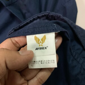 May include: Close-up of a navy blue garment with a white label. The label features the brand name AVIREX and a gold emblem of wings and a star. Japanese text is also visible on the label.
