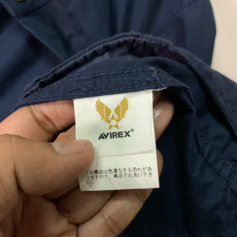 May include: Close-up of a navy blue garment with a white label. The label features the brand name AVIREX and a gold emblem of wings and a star. Japanese text is also visible on the label.