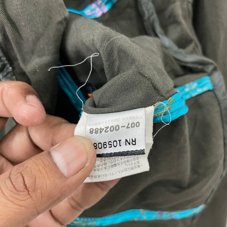 May include: Close-up of a dark green garment tag with the text "RN 105908 007-002488" printed on it. The tag is attached to a garment with a blue and white floral pattern.