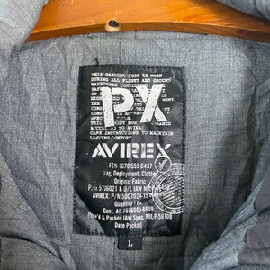 Pode incluir: Close-up de uma etiqueta preta Avirex numa jaqueta cinza. A etiqueta apresenta o texto "PX" em letras brancas grandes, juntamente com detalhes sobre a pe&ccedil;a de vestu&aacute;rio, incluindo a marca, informa&ccedil;&otilde;es sobre o tecido e o tamanho "L".
