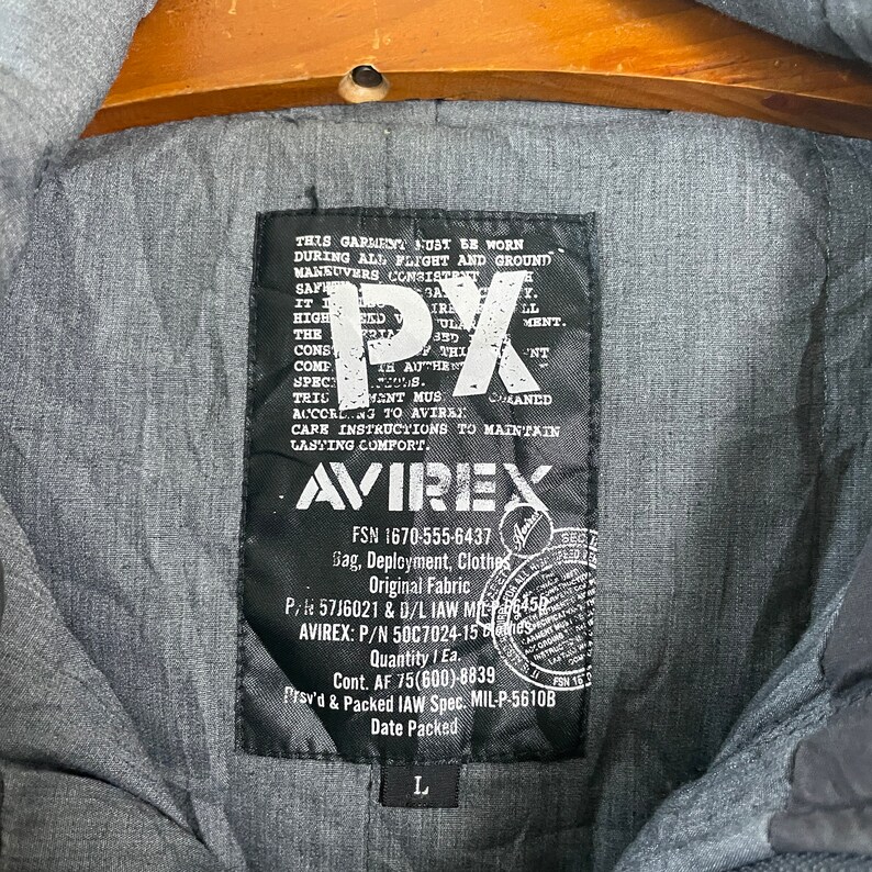 Pode incluir: Close-up de uma etiqueta preta Avirex numa jaqueta cinza. A etiqueta apresenta o texto "PX" em letras brancas grandes, juntamente com detalhes sobre a pe&ccedil;a de vestu&aacute;rio, incluindo a marca, informa&ccedil;&otilde;es sobre o tecido e o tamanho "L".