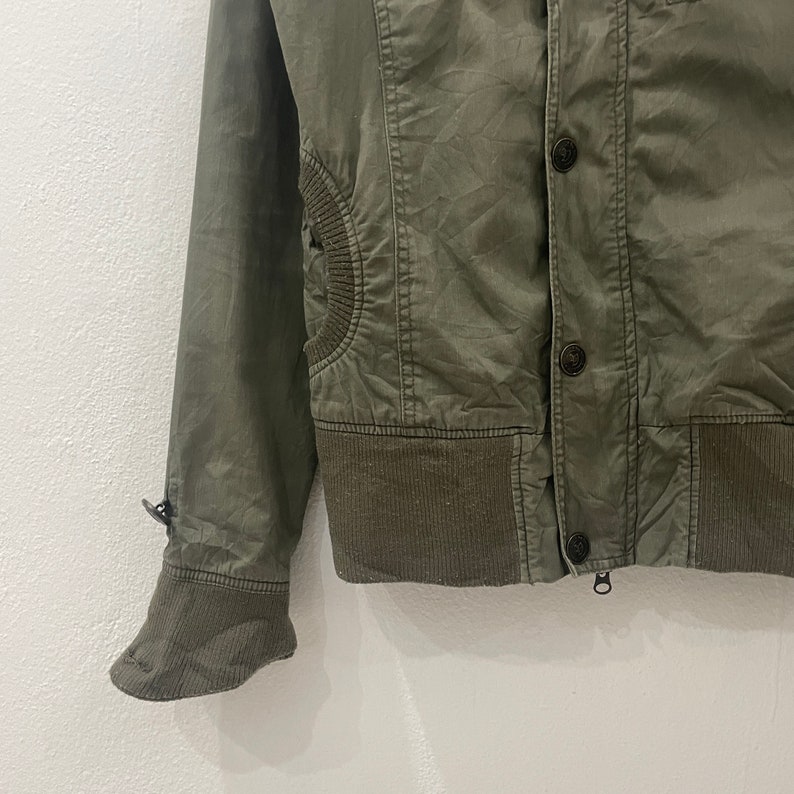 May include: A green utility jacket with a zipper closure and button accents. The jacket has a ribbed knit collar, cuffs, and waistband.
