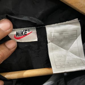 May include: Close-up of a black Nike jacket tag. The tag features the Nike logo in red and white, along with product information and size details. The tag reads "MADE IN MALAYSIA" and includes size "L".