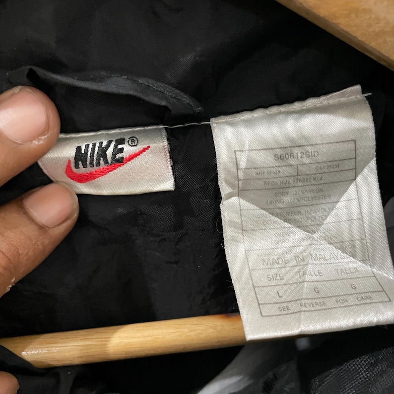 May include: Close-up of a black Nike jacket tag. The tag features the Nike logo in red and white, along with product information and size details. The tag reads "MADE IN MALAYSIA" and includes size "L".