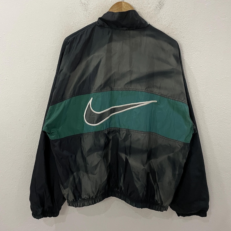 May include: Black and green Nike windbreaker jacket. The jacket features a large white Nike swoosh logo on the back, centered on a green stripe. The jacket is hanging on a wooden hanger.