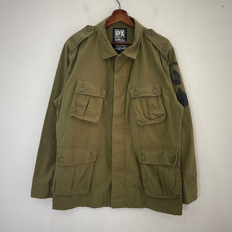 Op de afbeelding: Olijfgroene jas in militaire stijl met meerdere voorzakken en knoopsluitingen. De jas heeft een kraag en schouderstukken. Een zwarte afbeelding is zichtbaar op de linkermouw. De jas hangt aan een houten hanger.