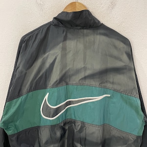 May include: Black and gray Nike windbreaker jacket with a green stripe across the chest. The Nike swoosh logo is white and prominently displayed. The jacket is hanging on a wooden hanger.