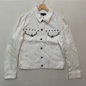 May include: White denim jacket with silver studs around the pockets and on the cuffs. The jacket has a button closure and a collared neckline.