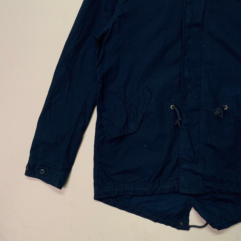 May include: A navy blue jacket with a drawstring waist and two front pockets. The jacket has buttoned cuffs and a small tassel on the drawstring. The jacket is made of a lightweight fabric.