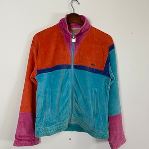Könnte beinhalten: Eine Vintage-Fila-Veloursjacke im Colorblock-Design. Die Jacke ist orange, blau und pink mit einem dunkelblauen Streifen über der Brust. Das Fila-Logo ist in Weiß auf die Brust gestickt.