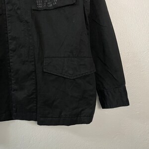 May include: Close-up of a black jacket with two front pockets and a chest pocket. The jacket has text on the chest pocket. The jacket is made of a durable fabric.