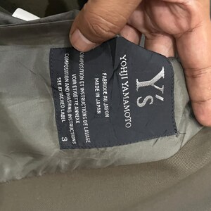 Puede incluir: Una etiqueta de prenda de color gris oscuro con el texto en blanco Y's Yohji Yamamoto, Fabrique au Japon, Made in Japan, y Composition et instructions de lavage, Voir etiquette annexe, Composition and washing instructions, See attached label, y el n&uacute;mero 3.