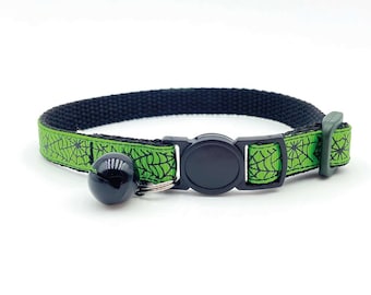 May include: A green cat collar with a black spiderweb pattern and a black bell. The collar has a black plastic buckle.