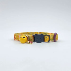 May include: A yellow and orange floral patterned cat collar with a black cat face buckle and a yellow bell charm.