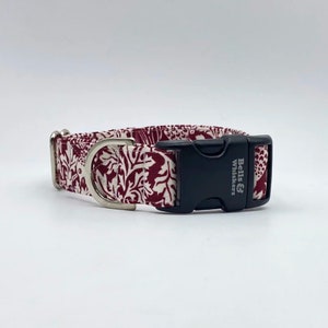 May include: A dog collar with a burgundy and white floral pattern. It has a black plastic buckle and a silver metal D-ring. The buckle is printed with the text "Bells & Whiskers".