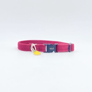 May include: A red fleece cat collar with a black buckle and a yellow pineapple charm. The buckle has the text "Belle & Whiskers" on it.
