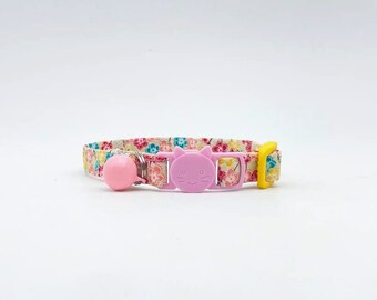 May include: A pink cat-shaped breakaway collar with a bell and a floral pattern. The collar is made of fabric and has a yellow buckle.