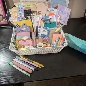 May include: A white plastic storage basket filled with various stationery items including notebooks, sticker sheets, pens, and small decorative erasers. A light blue zippered pencil case sits next to the basket, with five additional pens placed in front.