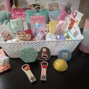 May include: A white plastic storage basket filled with various beauty and personal care items, including pink and teal pouches, hair combs, scrunchies, nail clippers with fruit charms, a rose-shaped compact, and a yellow bath bomb.