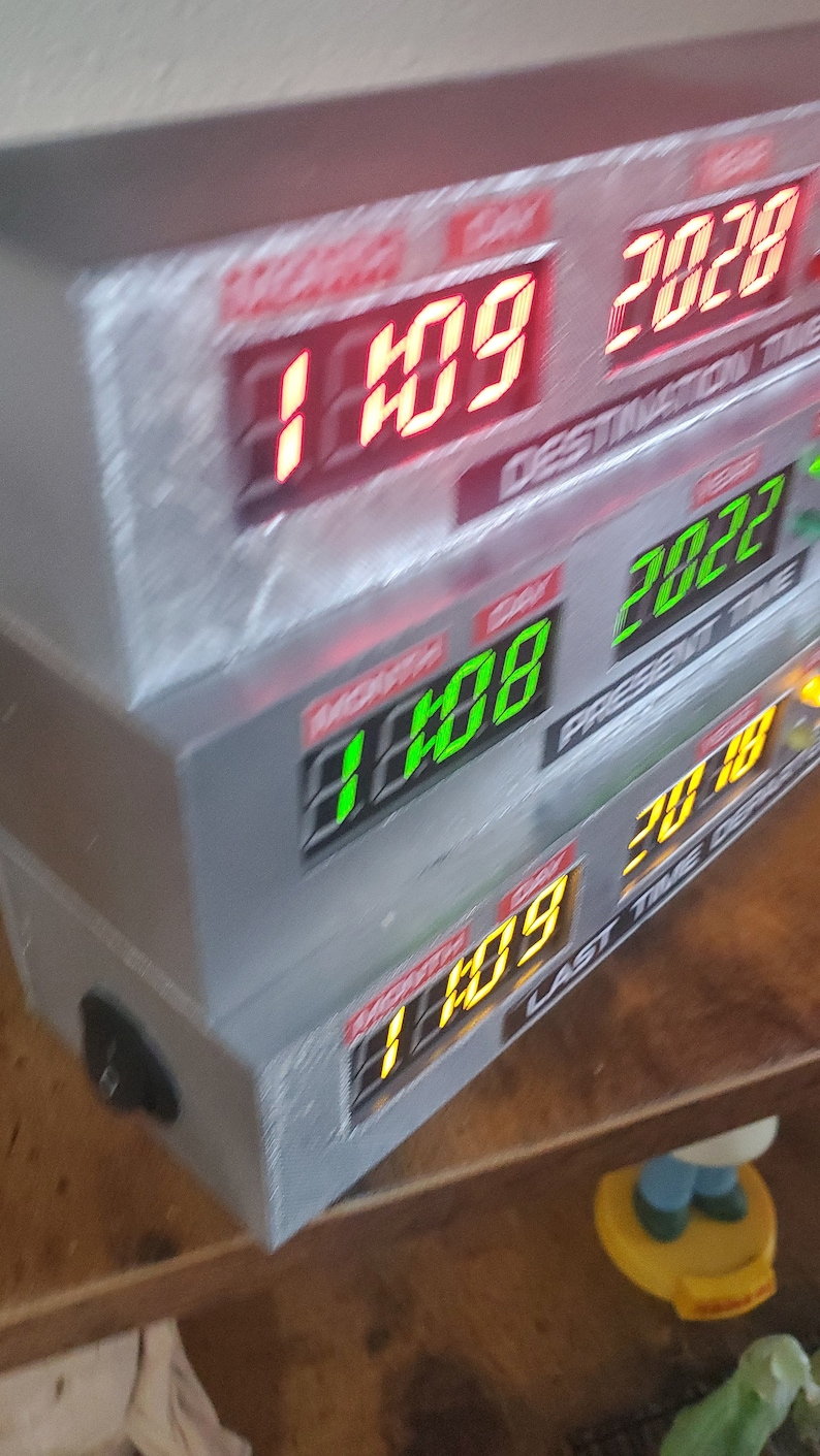 May include: A silver box with three digital displays showing the date and time. The top display shows "1:09" and "2028" in red, the middle display shows "1:08" and "2027" in green, and the bottom display shows "1:09" and "2018" in yellow. The text "DESTINATION TIME", "PRESENT TIME", and "LAST TIME" are visible on the displays.