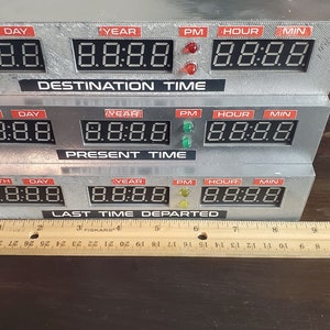 May include: A silver-colored time machine replica with three rows of digital displays. Each row has labels for month, day, year, PM, hour, and minute. The top row says "DESTINATION TIME", the middle row says "PRESENT TIME", and the bottom row says "LAST TIME DEPARTED".