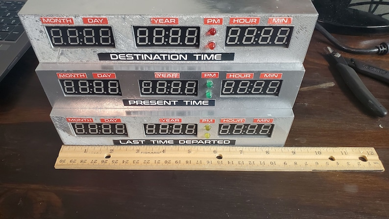 May include: A silver-colored time machine replica with three rows of digital displays. Each row has labels for month, day, year, PM, hour, and minute. The top row says "DESTINATION TIME", the middle row says "PRESENT TIME", and the bottom row says "LAST TIME DEPARTED".