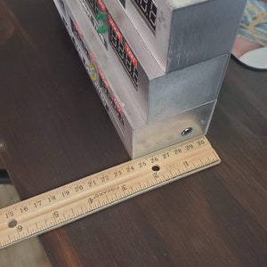 May include: A close-up of a wooden ruler measuring 30 inches, placed next to a stack of silver boxes with red and green lights and text on the front. The boxes are on a wooden surface.
