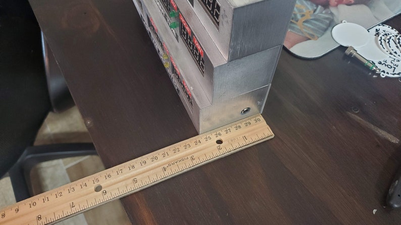 May include: A close-up of a wooden ruler measuring 30 inches, placed next to a stack of silver boxes with red and green lights and text on the front. The boxes are on a wooden surface.