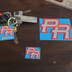 Könnte beinhalten: Drei blaue und weiße Schlüsselanhänger mit den Buchstaben "PR" in Rot. Der größte Schlüsselanhänger ist flach und rechteckig. Die beiden kleineren Schlüsselanhänger sind quadratisch. Der größte Schlüsselanhänger hat den Namen "Jaime" unter den Buchstaben gedruckt.