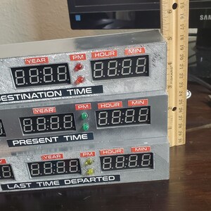 May include: A silver-colored time machine replica with three rows of digital displays. Each row has labels for month, day, year, PM, hour, and minute. The top row reads "DESTINATION TIME", the middle row reads "PRESENT TIME", and the bottom row reads "LAST TIME DEPARTED".