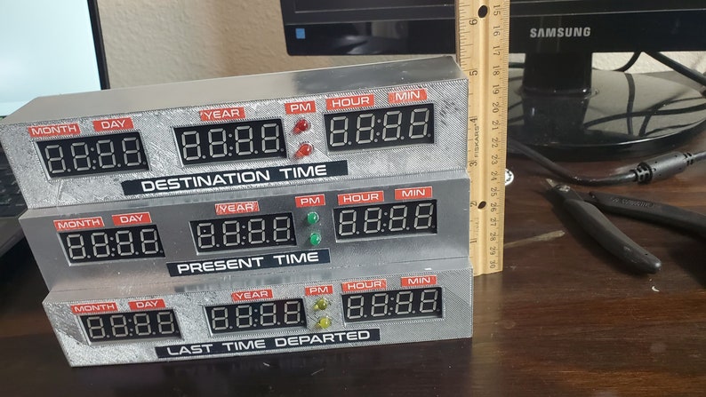 May include: A silver-colored time machine replica with three rows of digital displays. Each row has labels for month, day, year, PM, hour, and minute. The top row reads "DESTINATION TIME", the middle row reads "PRESENT TIME", and the bottom row reads "LAST TIME DEPARTED".