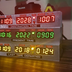 May include: A stack of three digital clocks with red, green, and yellow displays. The top clock displays "DESTINATION TIME" with the date "11:09 2028" and time "10:00". The middle clock displays "PRESENT TIME" with the date "10:16 2022" and time "09:09". The bottom clock displays "LAST TIME DEPARTED" with the date "1:09 2018" and time "01:24".