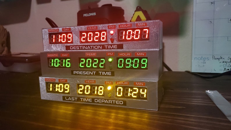 May include: A stack of three digital clocks with red, green, and yellow displays. The top clock displays "DESTINATION TIME" with the date "11:09 2028" and time "10:00". The middle clock displays "PRESENT TIME" with the date "10:16 2022" and time "09:09". The bottom clock displays "LAST TIME DEPARTED" with the date "1:09 2018" and time "01:24".