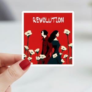 May include: A sticker with a red background featuring the word "REVOLUTION" in white. Two figures stand back-to-back, surrounded by white flowers. One figure wears a dark coat, the other a dark dress.