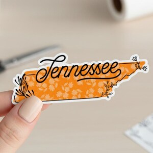May include: A sticker in the shape of Tennessee with the state's name in cursive black script. The state outline is orange with a floral pattern and black floral accents.