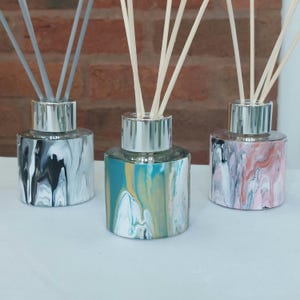 May include: Three reed diffusers with silver tops and patterned glass bases. One has black and white swirls, another teal and gold, and the third pink and white. Each has wooden reeds. The bases are approximately 7 cm in diameter.