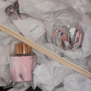May include: A gift box filled with white tissue paper containing a pink and grey marbled glass diffuser bottle with a gold lid and wooden diffuser sticks, and a pink and grey marbled glass candle holder. The box has a black label with the text "A+ glass gifts".