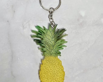 May include: A yellow pineapple keychain with a green top. The keychain is made of a clear material and has a silver key ring. The pineapple design is detailed and textured, giving it a realistic appearance.