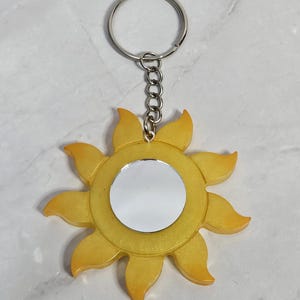 Könnte beinhalten: Ein gelber, sonnenförmiger Schlüsselanhänger mit einem silbernen Schlüsselring und einer Kette. Die Sonne hat eine verspiegelte Mitte und blütenblattartige Ränder. Ein fröhliches Accessoire.