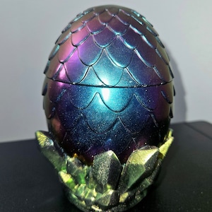 以下が含まれることがあります： 装飾的な、虹色のドラゴンの卵型の容器。卵は紫、青、緑の色合いが混ざり合い、鱗のような模様が施されています。緑と金の結晶の基盤の上に置かれています。容器は開くように設計されています。
