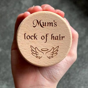 May include: A round, light-colored wooden keepsake box lid with the words "Mum's lock of hair" engraved in a decorative font. Below the text is an engraved image of angel wings with a halo. The lid is held in a hand.