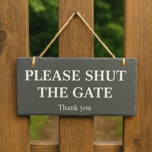 May include: A rectangular slate sign with the text "PLEASE SHUT THE GATE Thank you" in white lettering. The sign is suspended by a natural twine cord and hangs on a wooden fence.