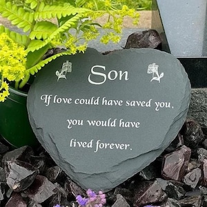 Peut inclure: Une pierre tombale en forme de cœur, grise, avec le mot "Son" et l'inscription "If love could have saved you, you would have lived forever." La pierre est entourée de roches sombres et de verdure.