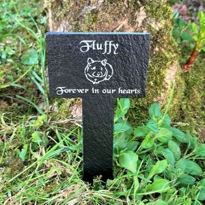 Op de afbeelding: Een zwarte leisteen gedenksteen voor huisdieren met de naam "Fluffy", een hamsterillustratie en de tekst "Forever in our hearts" in witte letters. De steen staat in een tuin met groen gras en bladeren, vlakbij een boomstam.