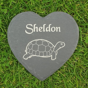 Pode incluir: Uma pedra de jardim em ardósia cinzenta em forma de coração com o nome "Sheldon" em escrita branca sobre um desenho de contorno branco de uma tartaruga. A pedra é colocada sobre um fundo de relva verde.